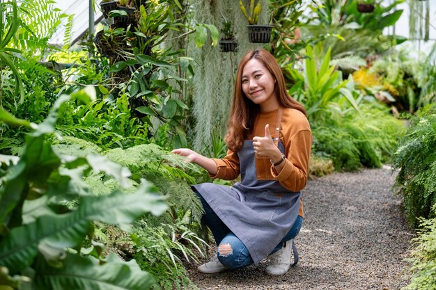 Un giovane giardiniere femminile asiatico che fa e mostra i pollici in su segno della mano mentre si prende cura delle piante nel giardino