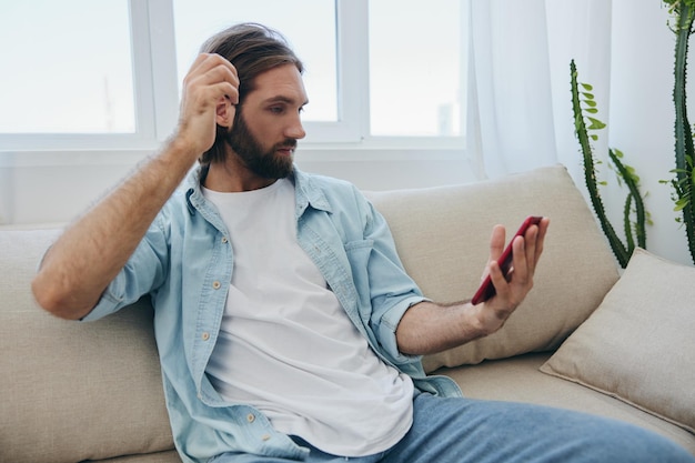 Un giovane guarda con rabbia e tristezza lo schermo del telefono a casa seduto sul divano Cattive notizie e sfortuna sul lavoro Foto di alta qualità