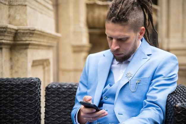 Un giovane hipster con i dreadlocks in un abito blu siede in un ristorante di strada guardando uno smartphone triste e sconvolto