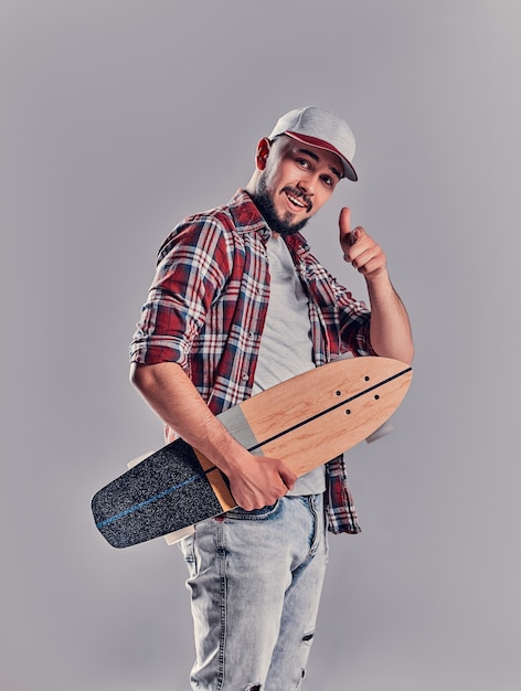 Un giovane hipster con la barba in camicia a quadri e berretto punta il dito contro qualcuno con un longboard