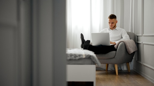 Un giovane in camicia lavora al laptop in camera da letto su una sedia con le gambe sul letto