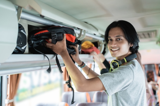 Un giovane in cuffia sorride guardando la telecamera quando mette una borsa su uno scaffale mentre si trova sull'autobus