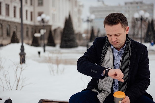 Un giovane in giacca e abito siede su una panchina in un parco cittadino e guarda l'orologio con una tazza di caffè