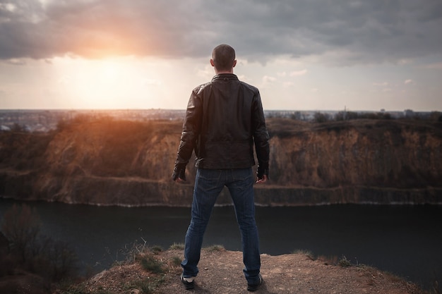 Un giovane in jeans e giacca di pelle marrone si trova sul bordo della vetta e guarda il lago al tramonto