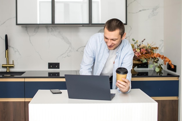 Un giovane in piedi in cucina sta lavorando su un laptop e tiene in mano una tazza usa e getta