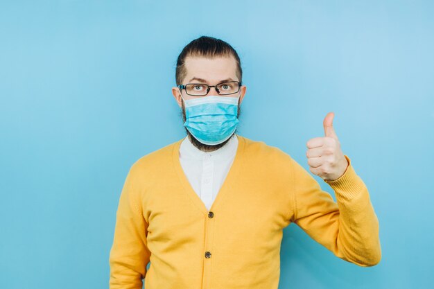 Un giovane in una mascherina medica. Un uomo in un cardigan giallo mostra un gesto della mano OK. Battiamo il coronavirus. L'epidemia di COVID-2019. Ragazzo positivo.