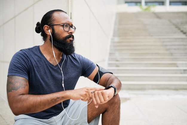 Un giovane indiano serio con la barba che ascolta l'audio negli auricolari e conta le pulsazioni con l'orologio fitness dopo aver fatto jogging