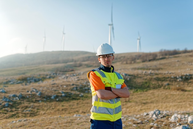 Un giovane ingegnere con le braccia incrociate ispeziona un campo di turbine eoliche mostrando fiducia e