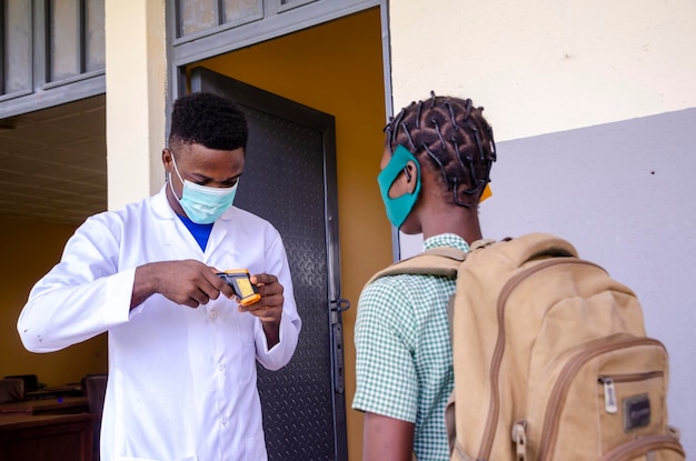 Un giovane insegnante di classe africano bello che tiene come termometro per scansionare la temperatura del suo studente prima di entrare in classe