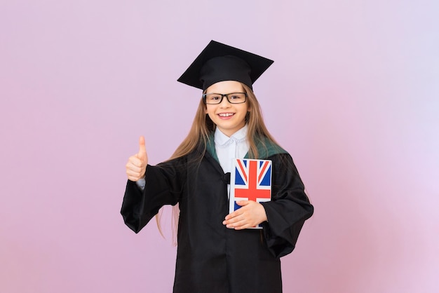 Un giovane laureato con un impermeabile cerimoniale e un cappello di laurea quadrato tiene un libro in lingua inglese e mostra un gesto della mano va tutto bene una scolaretta su uno sfondo isolato