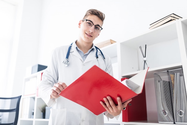 Un giovane medico in un abito bianco in piedi vicino al rack e in possesso di una cartella con documenti.