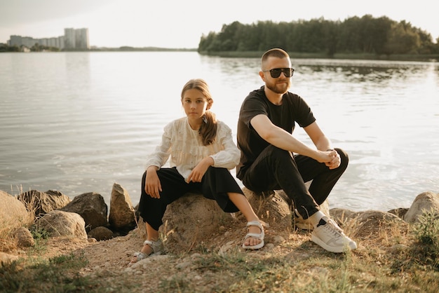 Un giovane padre con barba e occhiali da sole è seduto sui sassi con la figlia sulla costa del lago la sera. Famiglia monoparentale in vacanza.