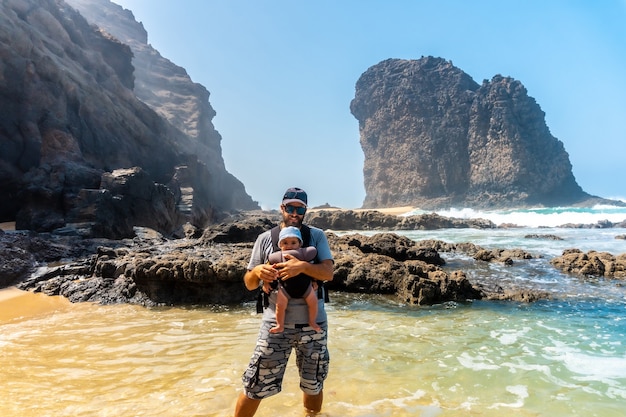 Un giovane padre con il suo bambino nello zaino nel Roque del Moro della spiaggia di Cofete del parco naturale di Jandia, Barlovento, a sud di Fuerteventura, Isole Canarie. Spagna