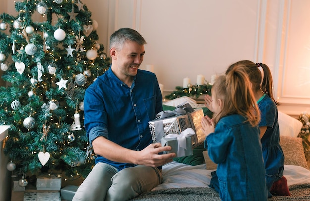 Un giovane padre fa regali di Natale alle sue figlie sorridenti vicino a un albero di Natale decorato