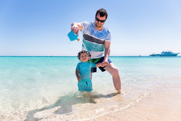 Un giovane padre sta giocando sulla spiaggia con il suo figlioletto e lo innaffia dall'annaffiatoio. Ragazzino che ride in riva al mare.