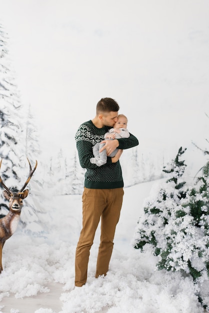 Un giovane papà tiene in braccio un figlio neonato in una zona fotografica in stile foresta invernale con un cervo e papà bacia suo figlio