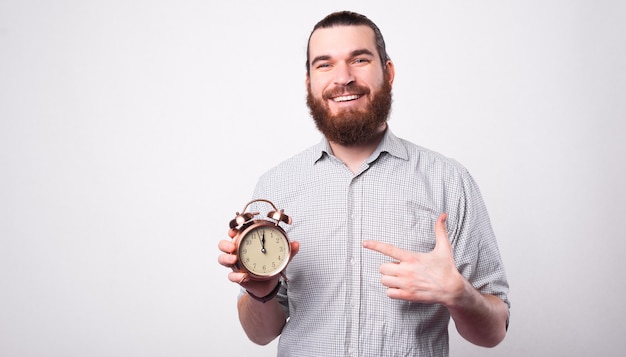 Un giovane positivo sorride alla telecamera e tiene un orologio indicandolo vicino a un muro bianco