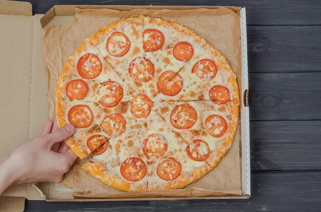 Un giovane prende una fetta di pizza con formaggio e pomodori in una scatola su uno sfondo di legno nero.