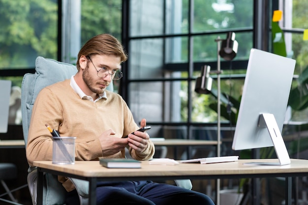 Un giovane programmatore libero professionista siede alla scrivania dell'ufficio e lavora al computer con in mano un telefono