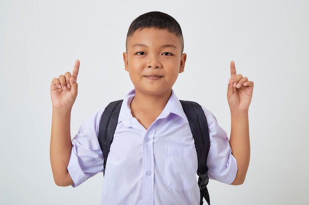 Un giovane ragazzo carino asiatico in piedi in un'uniforme scolastica thailandese con una borsa zaino e un libro su uno striscione bianco sullo sfondo