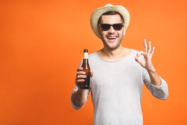 Un giovane ragazzo con un cappello e occhiali da sole isolati su uno sfondo arancione tiene una bottiglia di birra