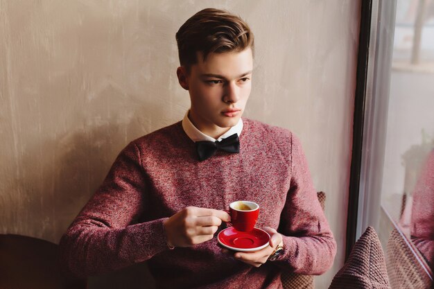 Un giovane ragazzo con un papillon sogna il futuro e beve caffè da una tazza rossa