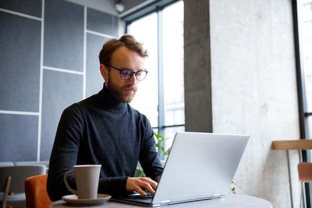 Un giovane ragazzo dai capelli rossi un programmatore o un imprenditore con gli occhiali lavora in un caffè alla moda dietro un laptop Il libero professionista lavora da remoto Comunicazione online Piccola impresa