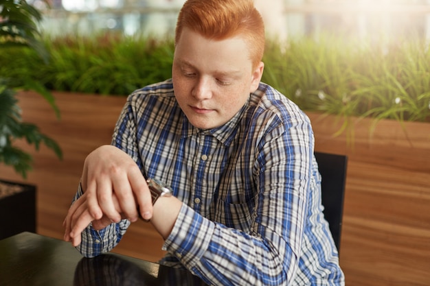 Un giovane ragazzo elegante con i capelli rossi e le lentiggini che indossa una camicia a quadri guardando il suo orologio in attesa dell'incontro nella caffetteria