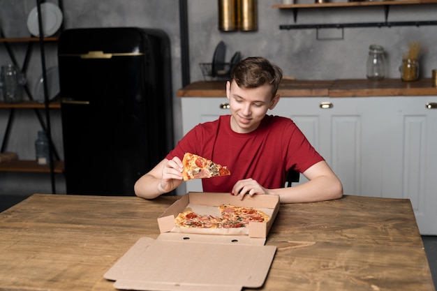 Un giovane si siede a un tavolo di legno con una pizza in una scatola di cartone e tiene in mano un pezzo di pizza