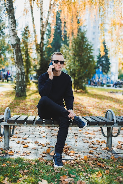 Un giovane solitario con un maglione nero siede nel parco su una panchina e parla al telefono Una calda giornata autunnale