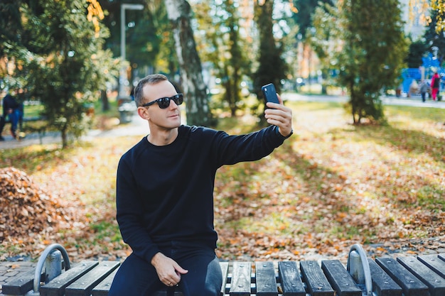 Un giovane solitario con un maglione nero siede nel parco su una panchina e parla al telefono Una calda giornata autunnale