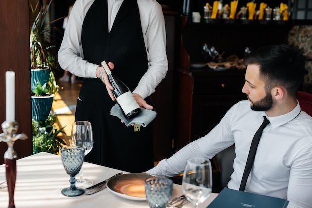 Un giovane sommelier in elegante divisa fa una dimostrazione e offre al cliente un buon vino nel ristorante Servizio clienti Allestimento tavola in un raffinato ristorante