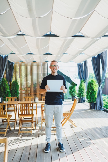 Un giovane sorridente si trova su una terrazza estiva in un bar con un laptop in mano Lavora come libero professionista online Lavoro estivo remoto online