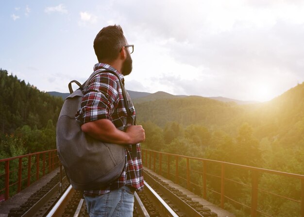 Un giovane turista con barba e occhiali in una camicia a quadri con uno zaino si trova sui binari della ferrovia e guarda in lontananza le montagne