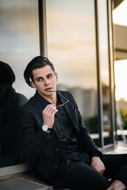 Un giovane uomo alla moda si trova vicino a un edificio a specchio durante il tramonto. Moda