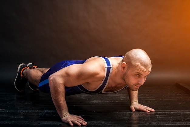 Un giovane uomo atletico in blu wrestling difficile e pantaloncini blu fa flessioni su uno sfondo isolato dai capelli neri in uno studio fotografico