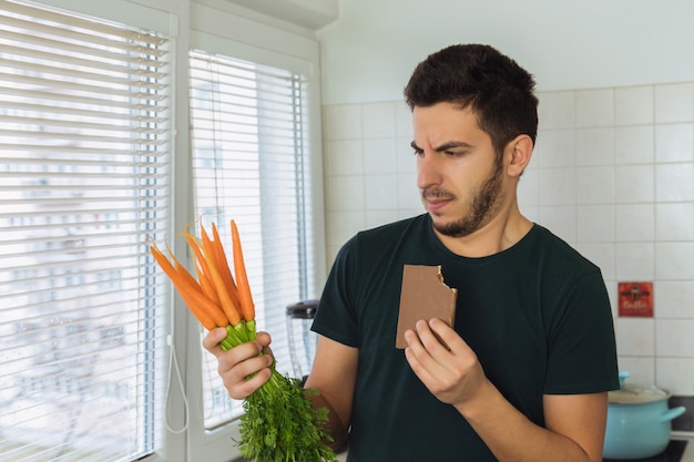 Un giovane uomo bruna guarda una carota con disprezzo e disgusto. Una persona non conduce uno stile di vita sano, vuole mangiare dolci e non cibi sani.