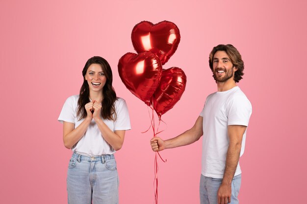 Un giovane uomo caucasico felice dà palloncini gonfiabili a forma di cuore rosso a una donna felice e scioccata
