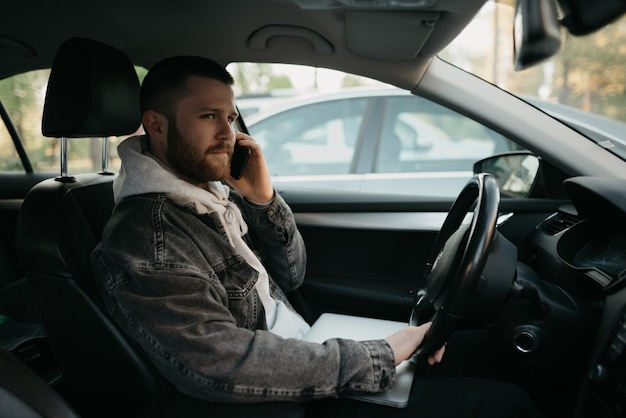 Un giovane uomo con la barba con il suo laptop e il suo smartphone all'interno di una macchina comfort