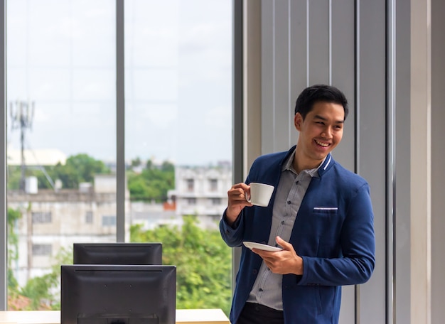 Un giovane uomo d'affari asiatico beve il caffè nella finestra dell'ufficio in un comodo