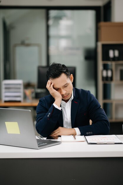 Un giovane uomo d'affari asiatico sovraccarico di lavoro, un impiegato che soffre di dolori al collo dopo una lunga giornata alla sua moderna scrivania, il concetto di sindrome dell'ufficio.