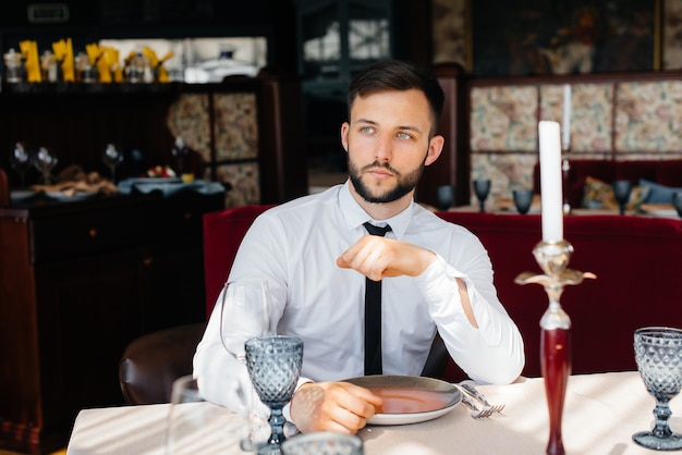 Un giovane uomo d'affari barbuto è seduto a un tavolo in un raffinato ristorante e aspetta il suo ordine. Servizio clienti nella ristorazione.