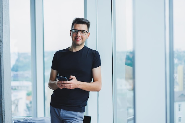 Un giovane uomo d'affari di successo con gli occhiali sta scrivendo un messaggio al telefono vicino alla finestra in un ufficio moderno e spazioso