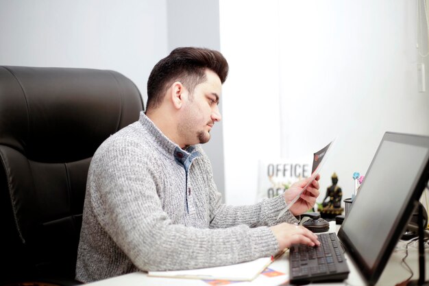 Un giovane uomo d'affari sta lavorando al computer tenendo la carta in mano.