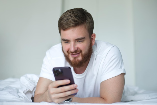 Un giovane uomo felice e barbuto sta usando il cellulare a casa nel letto nella camera da letto guardando lo schermo di