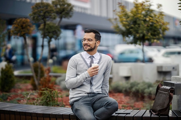 Un giovane uomo felice, intelligente e informale è seduto su una panchina fuori e si gode il suo caffè mattutino per andare