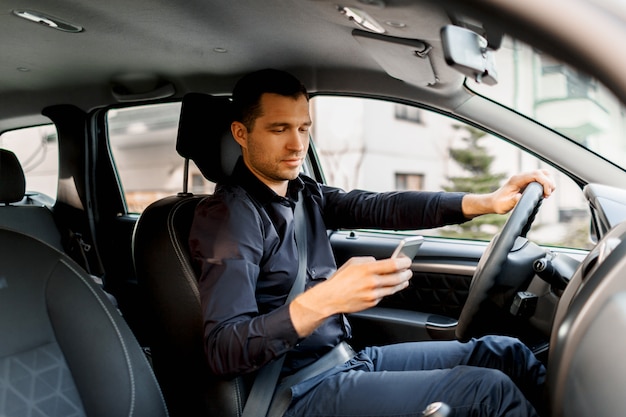 Un giovane uomo in una camicia scura alla guida della propria auto utilizza uno smartphone o un telefono. Concetto di multitasking