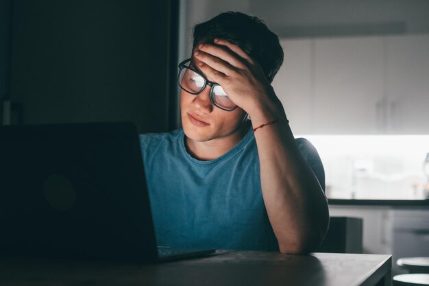 Un giovane uomo malato e stanco si sente male toccando gli occhi e il viso di notte dopo aver lavorato troppo Adolescente maschio con mal di testa che lavora