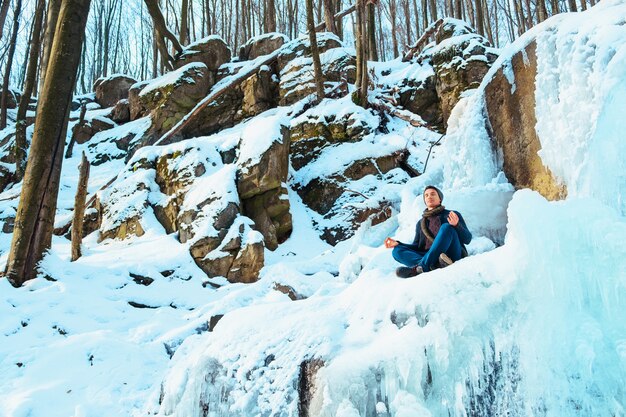 Un giovane uomo seduto nella neve cumulo di neve nella foresta e guardando il fiume d'inverno