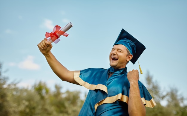 Un giovane uomo sorridente felice celebra il suo diploma di laurea e dottorato di ricerca nel cielo blu. festeggiare il concetto di laurea